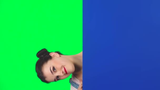 Ragazza guardando fuori da dietro cartello bianco blu sullo schermo verde in studio, rallentatore — Video Stock
