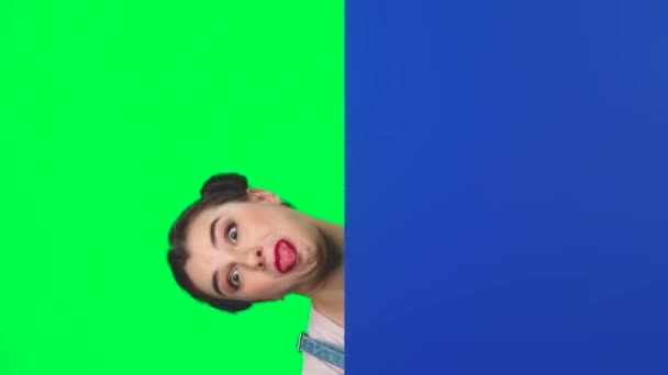 Смішна дівчина дивиться ззаду блакитного порожнього плакату на зеленому екрані на студії, повільний рух — стокове відео