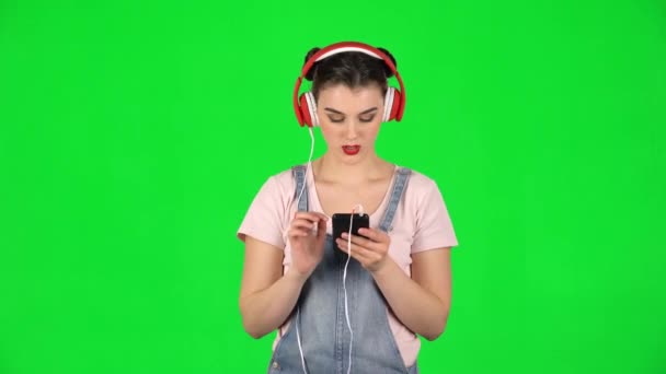 Menina em grandes fones de ouvido vermelhos escolhe música no telefone celular na tela verde — Vídeo de Stock