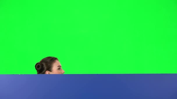 Girl looking out from behind blue blank placard on green screen at studio. The board is horizontal — Stock Video