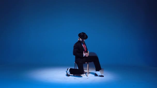 Un hombre elegante con un sombrero negro está bailando un baile erótico. Proyector sobre fondo azul . — Vídeos de Stock