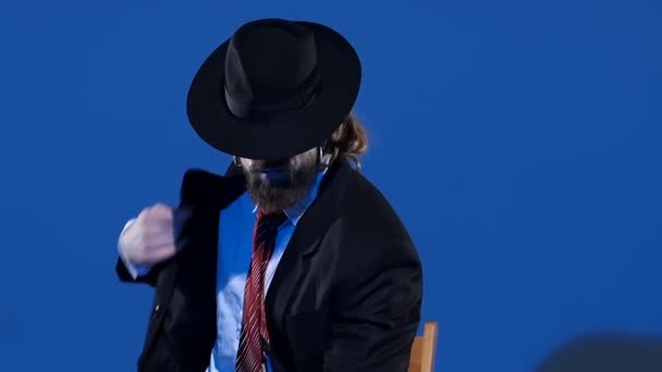 Un hombre elegante con un sombrero negro está bailando un baile erótico. Proyector sobre fondo azul. Primer plano, cámara lenta . — Vídeo de stock