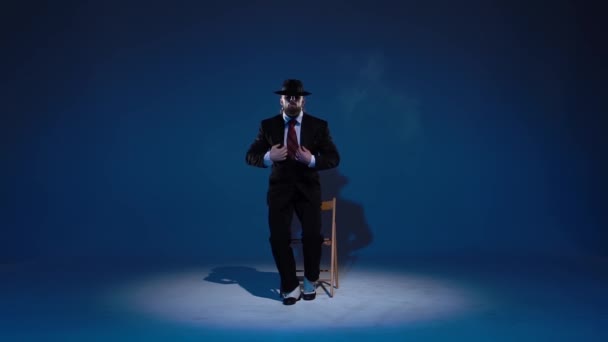 Элегантный мужчина в черной шляпе танцует эротический танец. Прожектор на синем фоне. Закройте, замедленная съемка . — стоковое видео