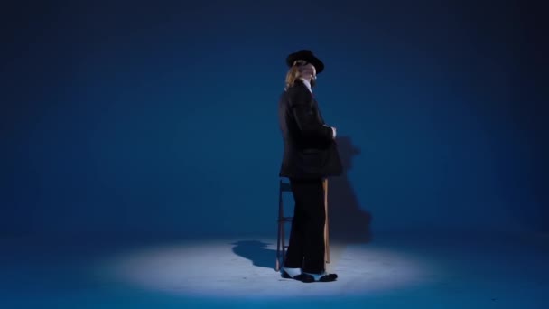 Elegant man in a black hat is dancing an erotic dance. Spotlight on a blue background. Close up, slow motion. — ストック動画