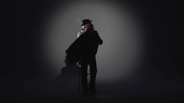 Un hombre elegante con un sombrero negro está bailando un baile erótico. Proyector sobre fondo negro. Primer plano, cámara lenta . — Vídeos de Stock