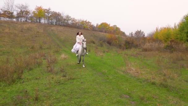 Довгошерста дівчина в білій сукні верхи на коні вздовж лісу — стокове відео