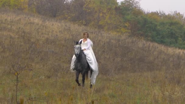 Όμορφη αρραβωνιαστικιά με λευκό φόρεμα ιππεύει άλογο κοντά στο ξύλο — Αρχείο Βίντεο