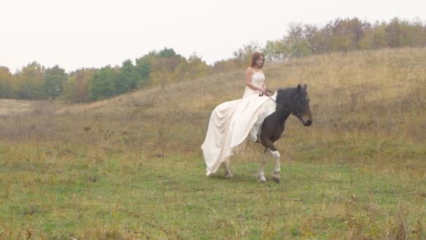 Menina de cabelos vermelhos em vestido poofy está montando cavalo ao longo do campo — Vídeo de Stock