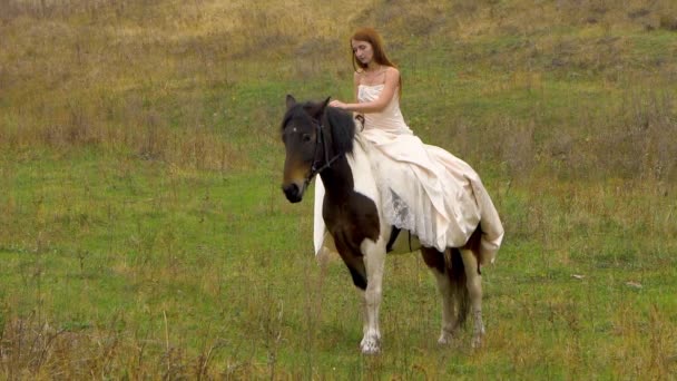 Рыжая женщина в пушистом платье едет верхом на лошади по лугу — стоковое видео