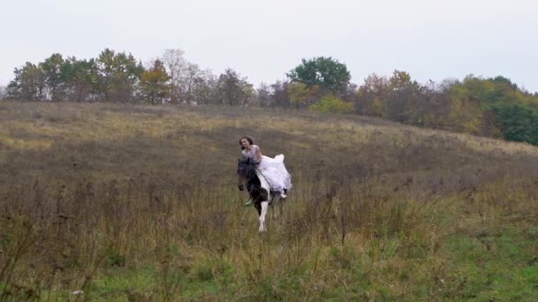 Langharige paardenvrouw in trouwjurk paardrijden galopperend paard langs veld — Stockvideo