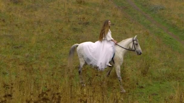 Long-haired fiancee in white wedding dress riding horse through valley — Stock Video