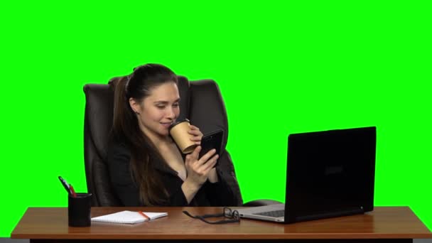 Business woman sitting at desk in leather chair writes message on cell phone and drink coffee. Green screen. Studio. Slow motion — ストック動画