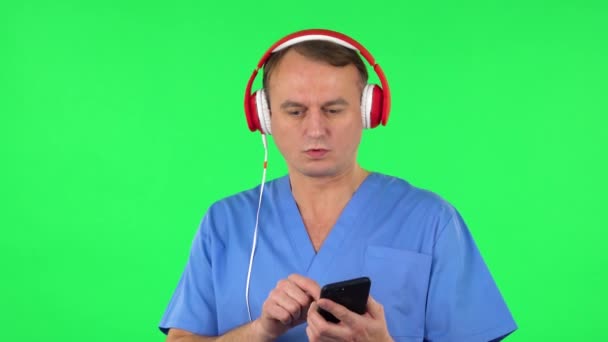 A nagy piros fülhallgatós orvos bekapcsolja a zenét a mobiltelefonnál. Zöld képernyő — Stock videók