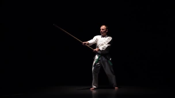 Боец Кендо на белом кимоно практикует боевое искусство с бамбуковым боккеном на черном фоне. Медленное движение — стоковое видео