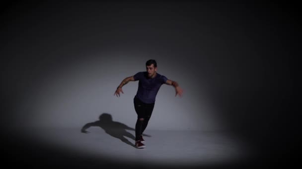 男はシンプルなグレーのTシャツとグレーのジーンズで暗い背景にブレークダンスを踊っています — ストック動画