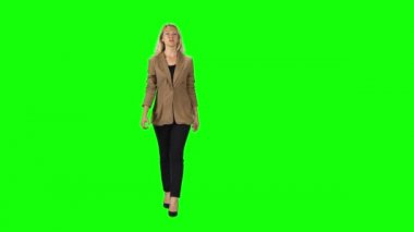 Blonde girl in a stylish brown jacket, black trousers and high-heeled shoes going against a green screen.