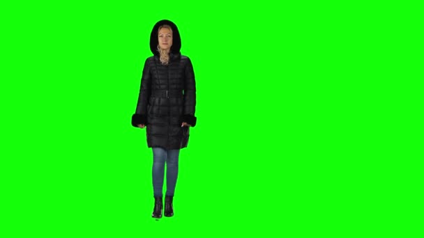 Blonde girl in a black winter coat with hood, jeans and boots going against a green screen. — ストック動画