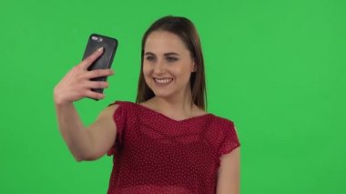 Portrait of tender girl in red dress is making selfie on mobile phone then looking photos. Green screen