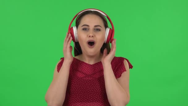 Portrait of tender girl in red dress is dancing and enjoying music in big red headphones. Green screen — Stock Video