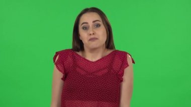 Portrait of tender girl in red dress is saying oops and shrugging and biting lips. Green screen