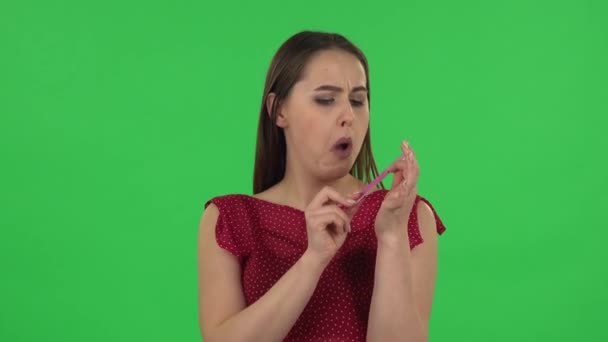 Portrait of tender girl in red dress is making herself manicure with pink nail file. Green screen — Stockvideo