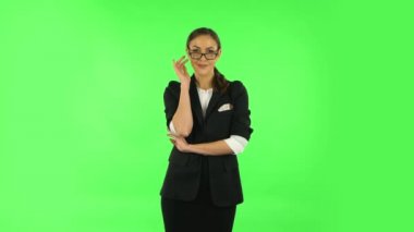 Esmer kız takım elbiseli, gözlüklerini düzeltiyor, kaşlarını kaldırıyor ve yeşil ekrana gülümsüyor.