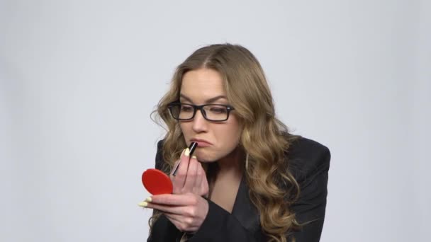 Όμορφη γυναίκα με γυαλιά ζωγραφίζει τα χείλη της κοιτάζοντας σε κόκκινο καθρέφτη σε γκρι φόντο — Αρχείο Βίντεο