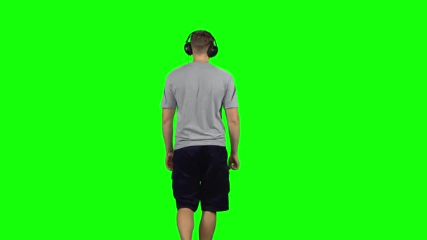 El hombre con auriculares grandes va, escuchando música y bailando en pantalla verde en el estudio. Vista trasera — Vídeos de Stock