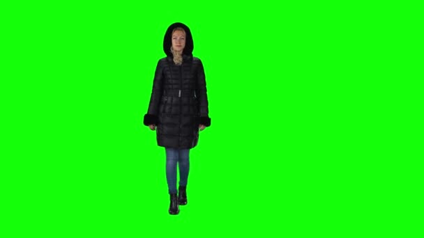 Blond tjej i svart vinterrock med huva, jeans och stövlar som går mot en grön skärm. Långsamma rörelser — Stockvideo