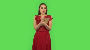 Tender girl in red dress is standing and thinking, then happy writing with pencil in notebook. Green screen