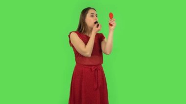 Tender girl in red dress is painting her lips looking in red mirror. Green screen