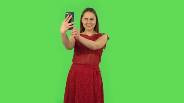 Нежная девушка в красном платье делает селфи на мобильном телефоне, а затем смотрит фотографии. Зеленый экран — стоковое видео