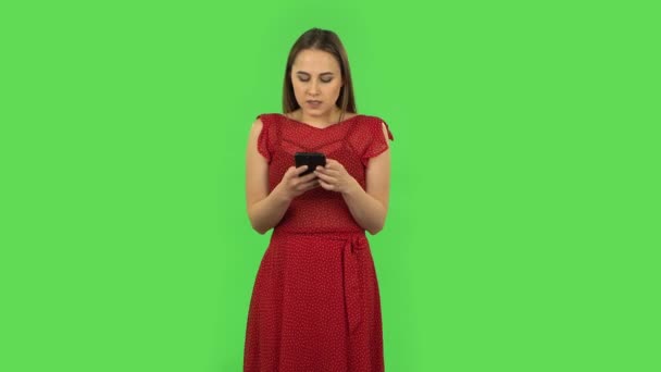 Tender girl in red dress is angrily texting on her phone. Green screen — ストック動画