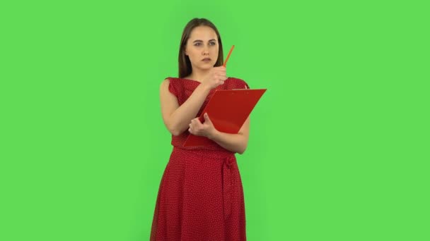 Tender girl in red dress fills papers in red folder with pensil. Green screen — Stockvideo