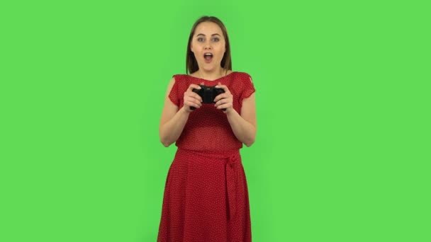 Tender girl in red dress is playing a video game using a wireless controller and rejoicing in victory. Green screen — Stock videók