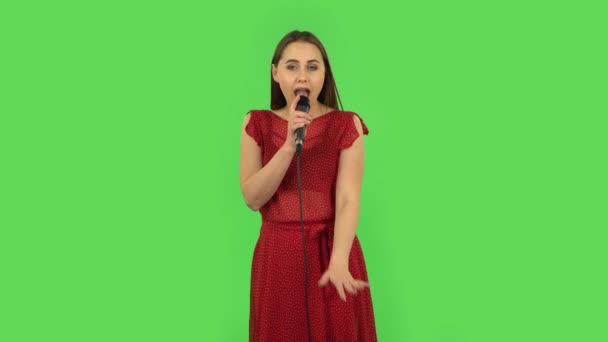 Нежная девушка в красном платье поет в микрофон и движется под ритм музыки. Зеленый экран — стоковое видео
