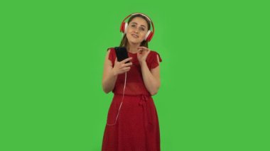 Tender girl in red dress is dancing and enjoying music in big red headphones. Green screen