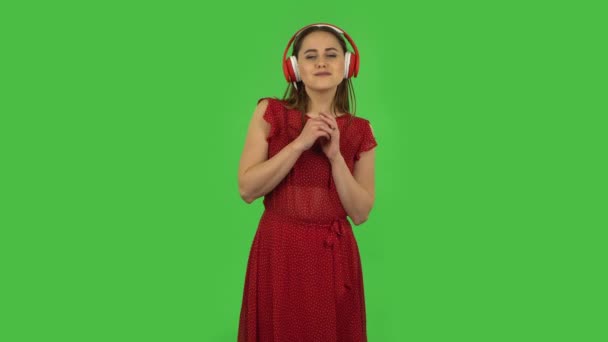 Нежная девушка в красном платье танцует и наслаждается музыкой в больших красных наушниках. Зеленый экран — стоковое видео