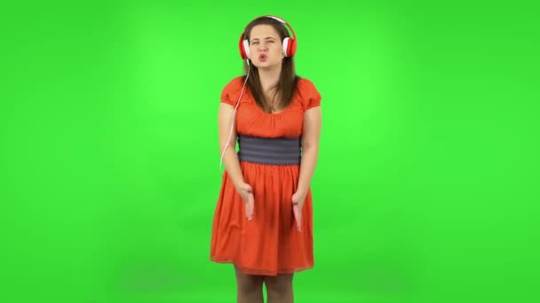 Cute girl jest taniec i cieszyć się muzyką w dużych czerwonych słuchawkach. Zielony ekran — Wideo stockowe