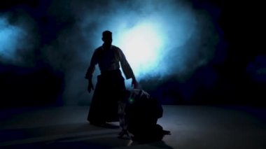 İki dövüşçü aikido tekniği uyguluyor. Karanlık stüdyoda duman ve ışık saçan ustaların siluetleri..