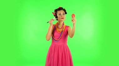 Girl with curlers on her head in a pink dress looking in red mirror and powdered her nose with big brush. Green screen