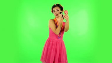 Girl with curlers on her head in a pink dress looking in red mirror and powdered her nose with big brush. Green screen