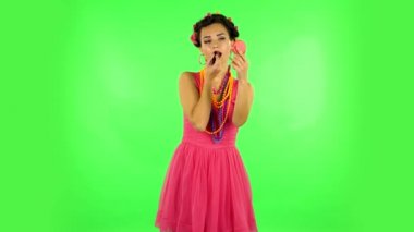 Girl with curlers on her head in a pink dress paints her lips looking in red mirror. Green screen