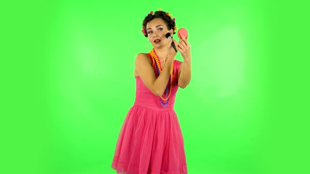 Girl with curlers on her head in a pink dress looking in red mirror and powdered her nose with big brush. Green screen — 图库视频影像