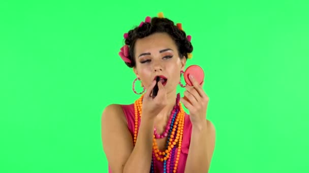Girl with curlers on her head in a pink dress looking in red mirror and paints her lips. Green screen — Αρχείο Βίντεο