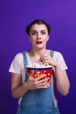 Elinde patlamış mısır kabıyla korku filmi izleyen güzel bir bayan mor arka planda izole edilmiş günlük kıyafetler giyiyor.