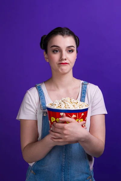 Chica bonita sosteniendo palomitas de maíz contenedor mirando a la cámara, aislado en el fondo púrpura —  Fotos de Stock