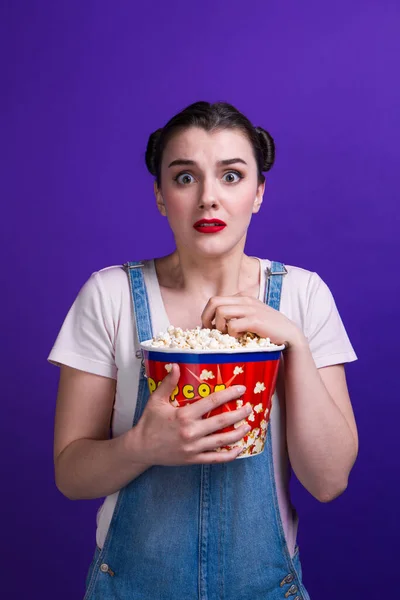 Mooie dame holding popcorn container kijken horror film dragen casual outfit geïsoleerd op paarse achtergrond — Stockfoto