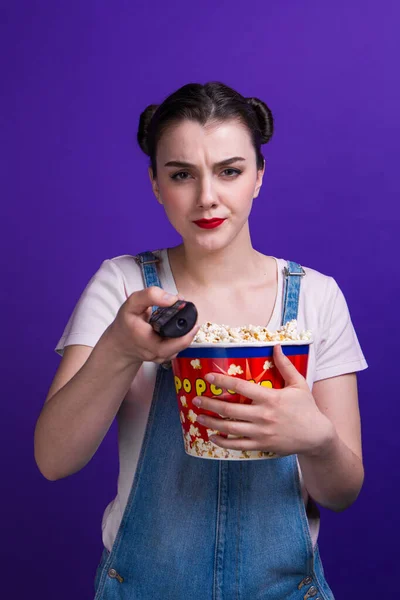 Meisje met grote popcorn box, staren verandering kanaal terwijl het kijken naar televisie met afstandsbediening geïsoleerd op paarse achtergrond — Stockfoto