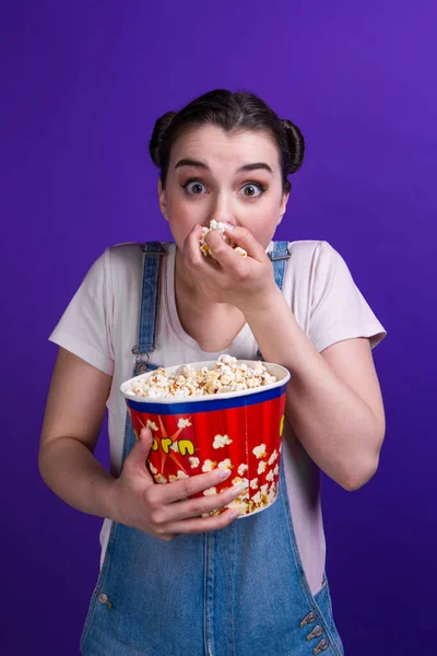 Girl with two buns watch tv show and eating popcorn bucket mouth open, thrill exciting plot, isolated blue background